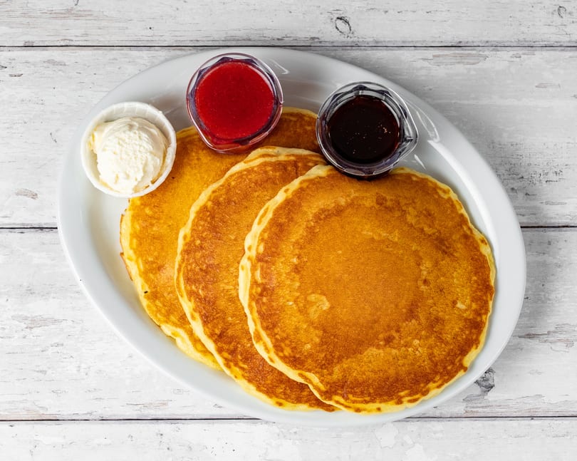Three golden pancakes on a white plate with butter, strawberry jam, and maple syrup.