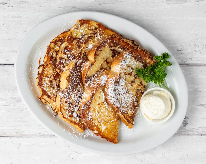Golden brown French toast slices dusted with powdered sugar, served with butter and parsley on a white plate.